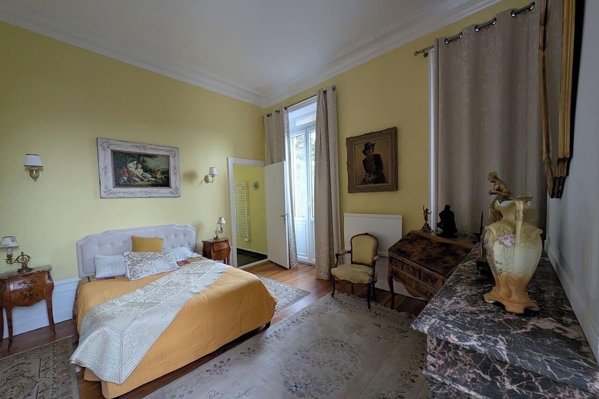 A bedroom with a bed, a desk, a chair, a vase, and a painting on the wall.