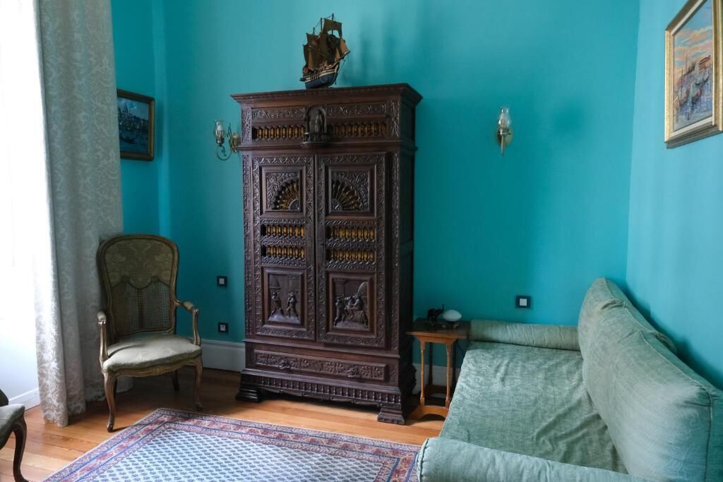 Una habitación con una pared verde azulado, un mueble de madera oscura con adornos, un sillón verde y una alfombra estampada.