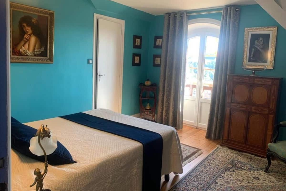 A bedroom with a bed, a door, a window with curtains, a small table, and a cabinet.
