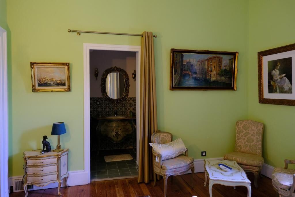A room with a wooden floor, a small table with a lamp, a chair, a bed with a headboard, and a painting on the wall.