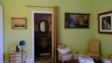 A room with a wooden floor, a small table with a lamp, a chair, a bed with a headboard, and a painting on the wall.