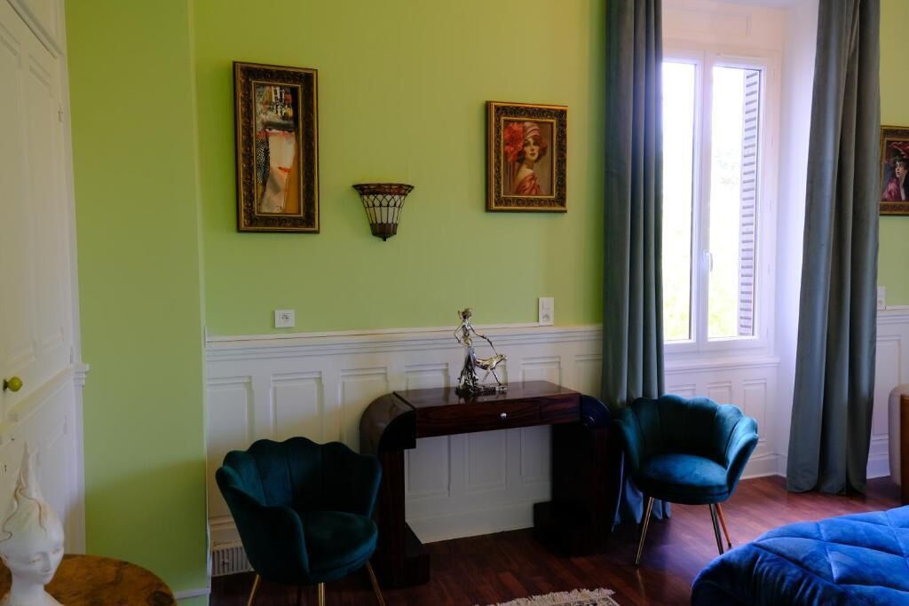 A room with a green wall, a desk with a lamp, two chairs, a bed, and a window with curtains.