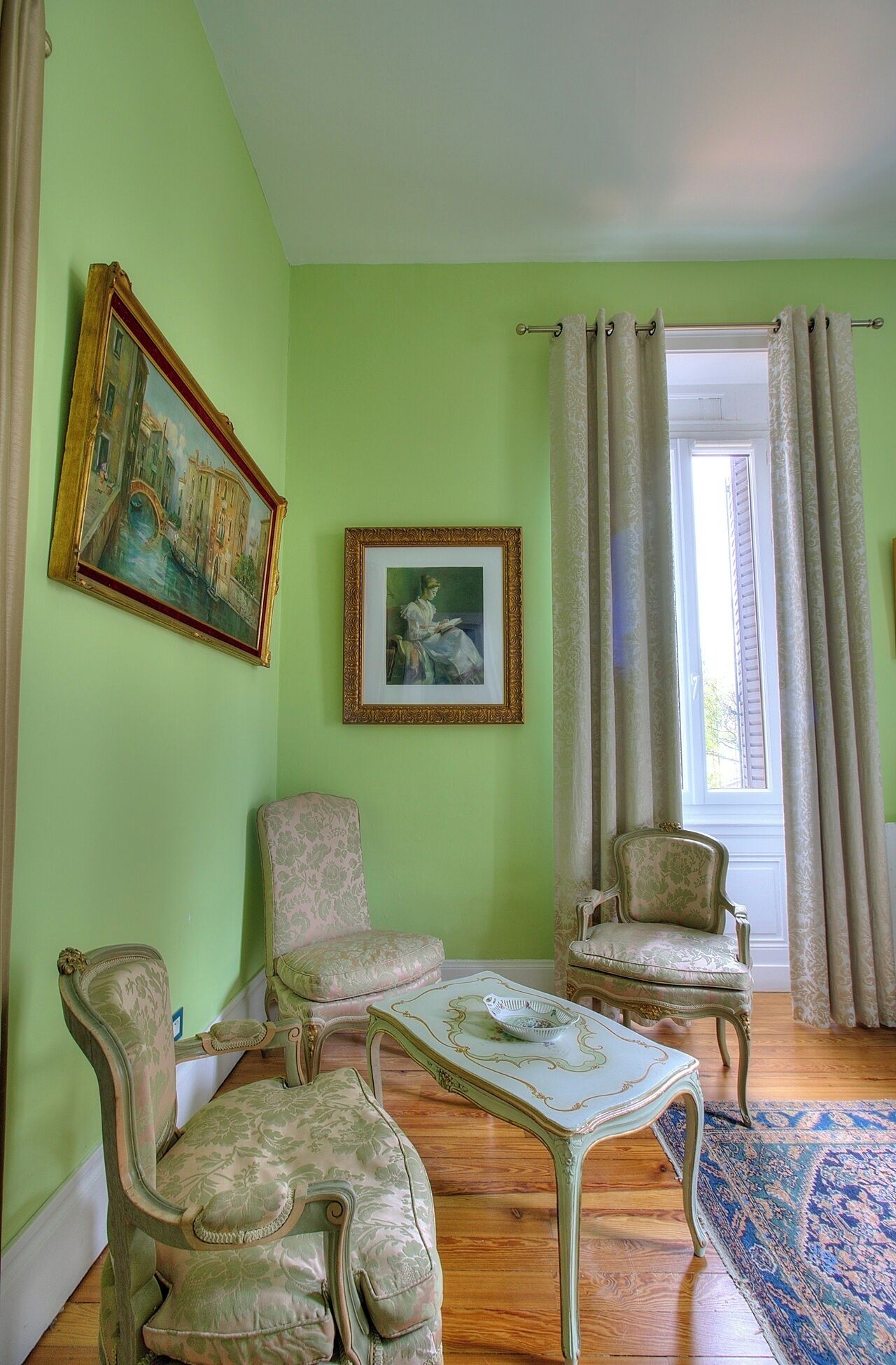 Una habitación con paredes verdes, dos sillas de estilo antiguo, una mesa pequeña y un cuadro representando una escena de un canal.