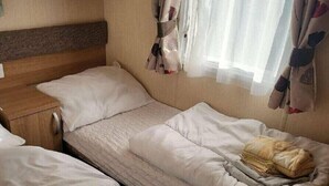 A single bed with a wooden headboard, a bedside table, and a window with curtains. - Weymouth Bay Holiday Park - Holiday Accommodation 15694 (Weymouth)