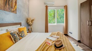 Ein Schlafzimmer mit Bett, Kissen, Decke, Hut, Fenster mit Vorhängen, einem Wandbild und einer Klimaanlage.