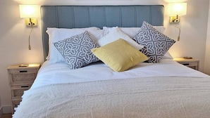 Ein Bett mit blauem Kopfteil, weißer Bettwäsche und gemusterten Kissen. Darüber hängt ein gerahmtes Bild, und an beiden Seiten befinden sich Wandleuchten.