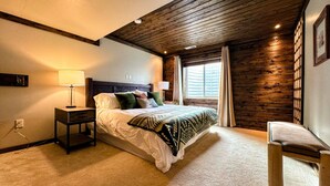 A bedroom with a bed, bedside tables, a nightstand, a window with curtains, and a wooden ceiling.