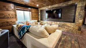 A cozy room with a stone wall, a mounted TV, a sofa with cushions, and a window with blinds.