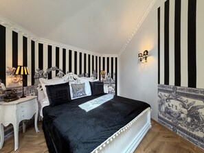 A bedroom with a black and white striped wall, a bed with white and black bedding, a nightstand with a lamp, and a decorative wall piece. - Chateau D'Amour Luxury Boutique (Bucharest)