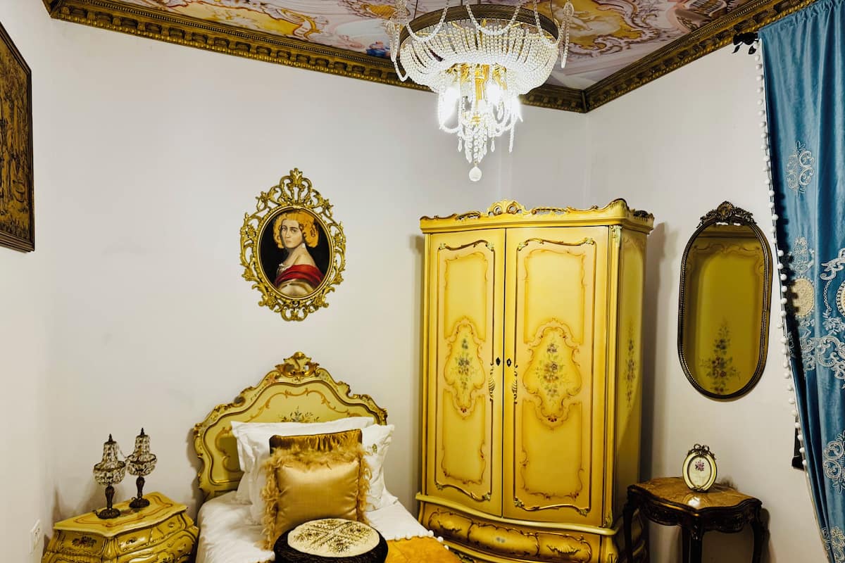 A room with a bed, a wardrobe, a chandelier, a mirror, and a picture on the wall.