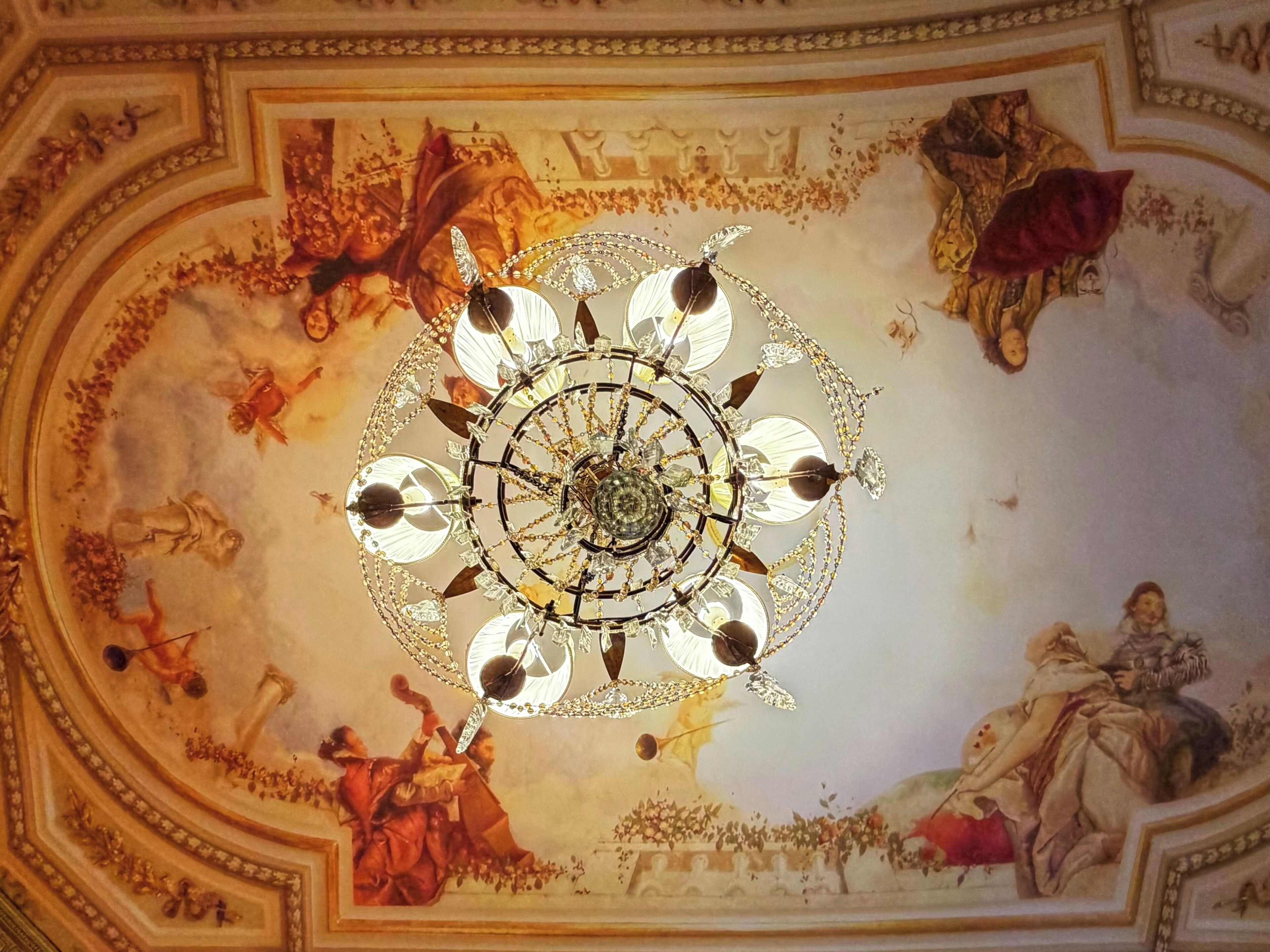Un techo con una lámpara central y frescos que representan escenas históricas.