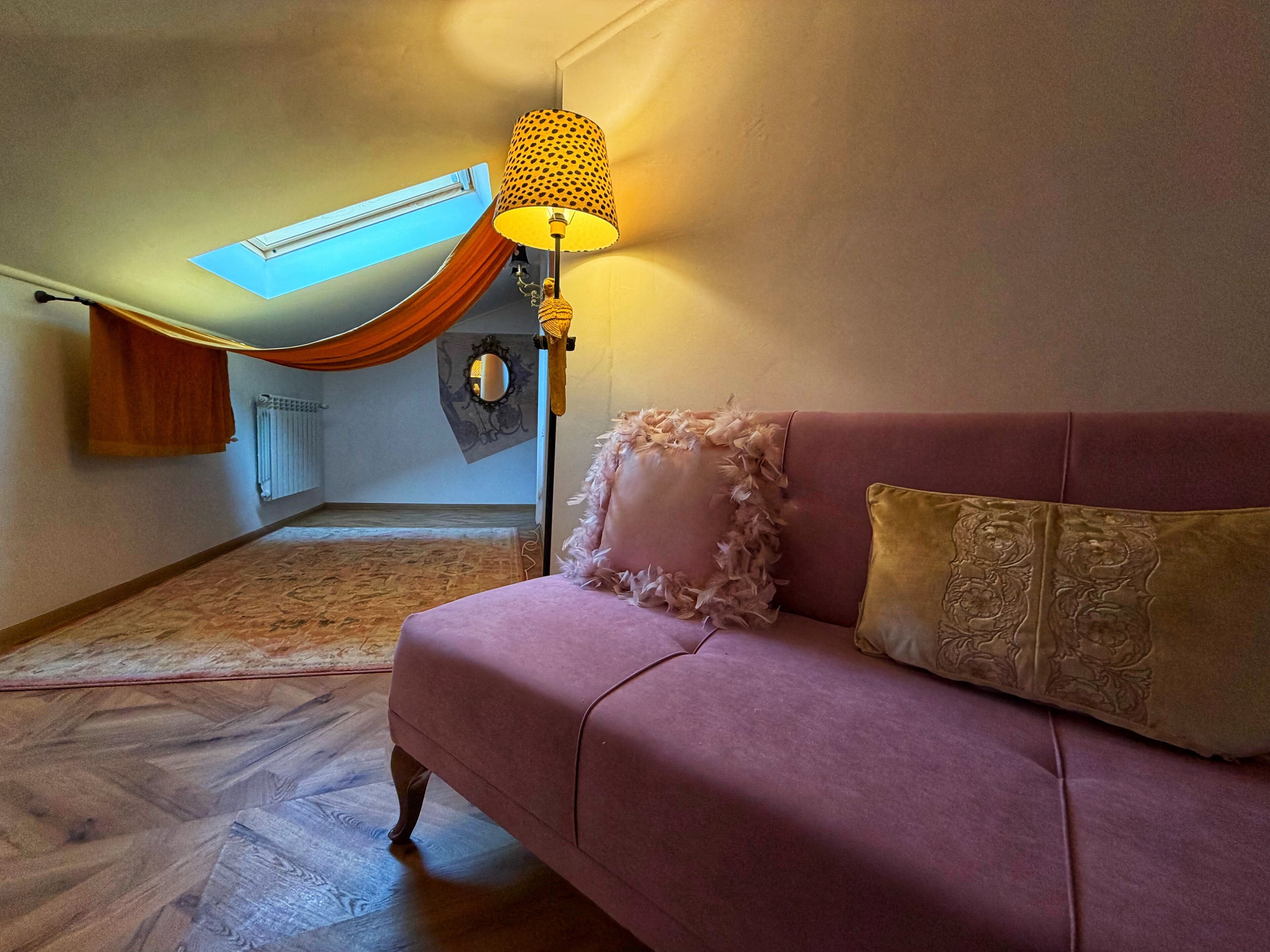 Una habitación con un sofá rosa, una lámpara, un espejo y una ventana con cortina.