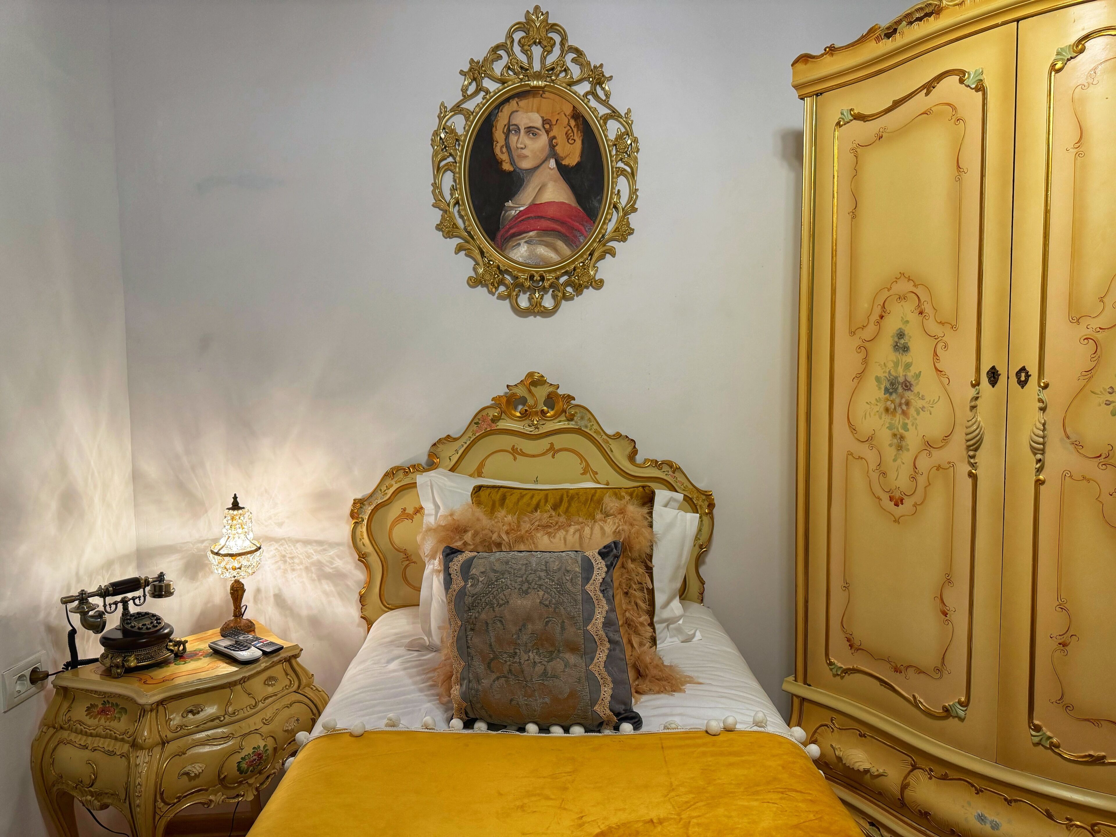 A room with a bed, a nightstand with an old-fashioned telephone, a lamp, a wardrobe, and a framed picture on the wall.