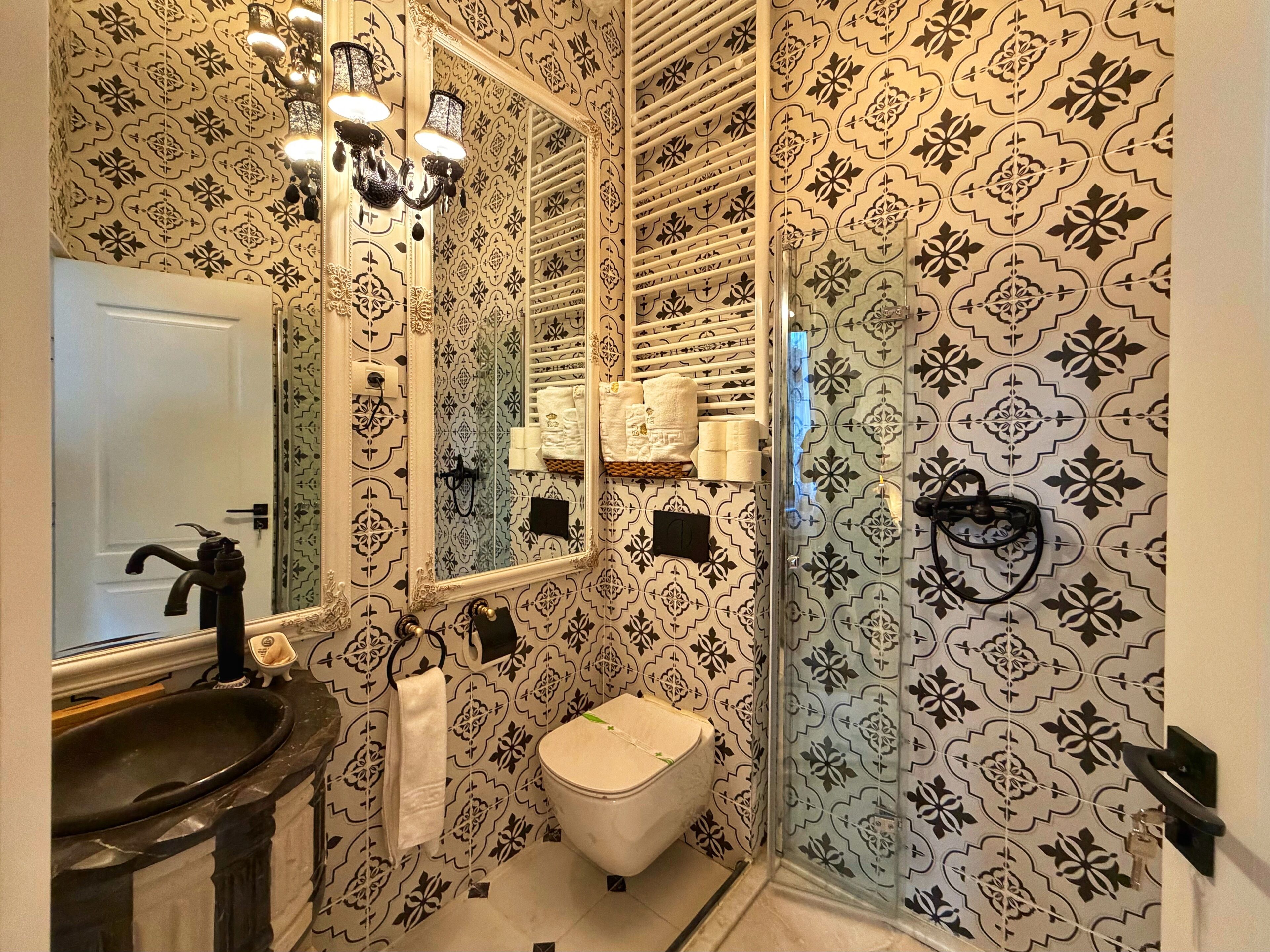 Un baño con lavamanos, inodoro y zona de ducha, con papel tapiz estampado y un espejo.