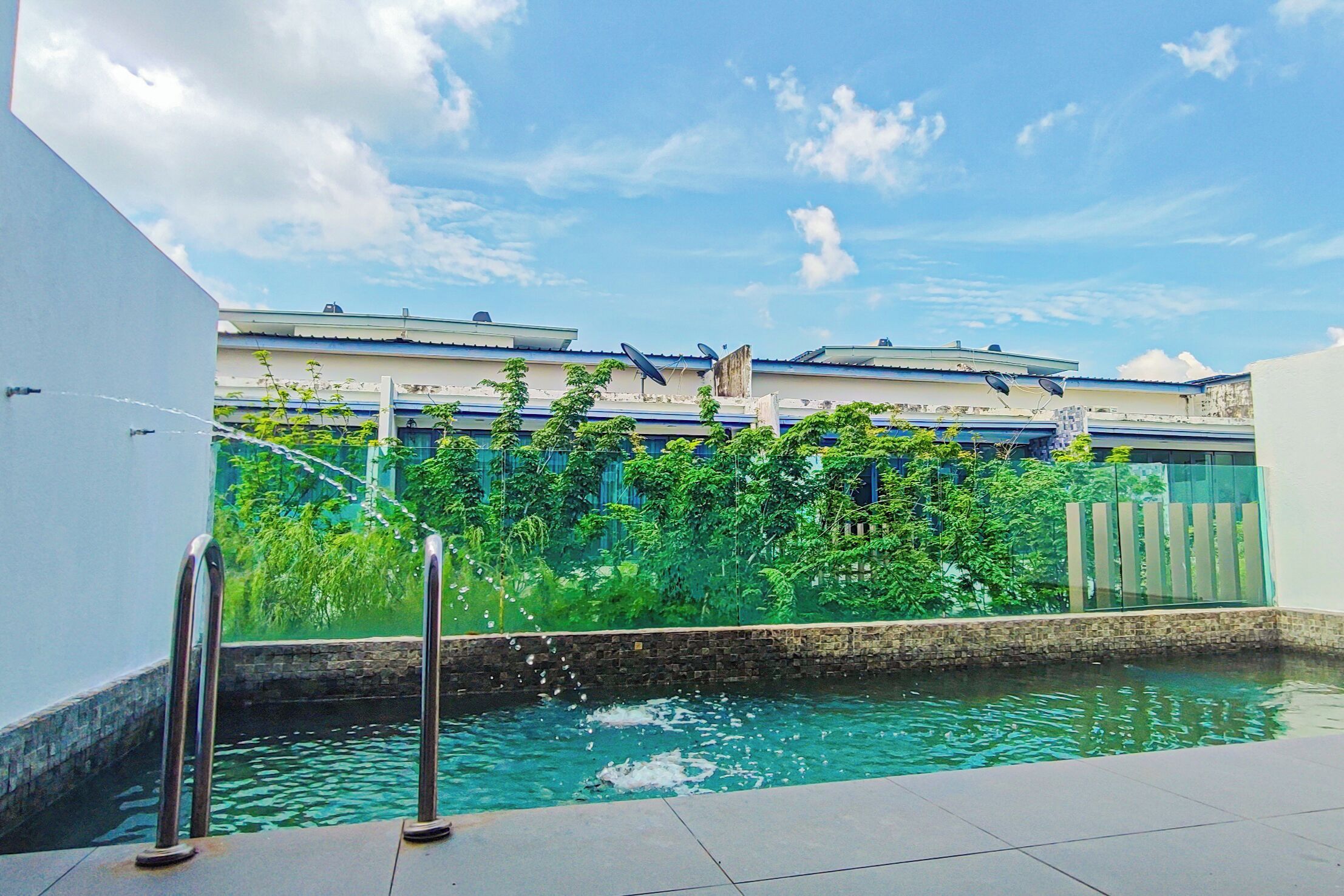 A swimming pool with a water fountain, surrounded by greenery and buildings in the background.