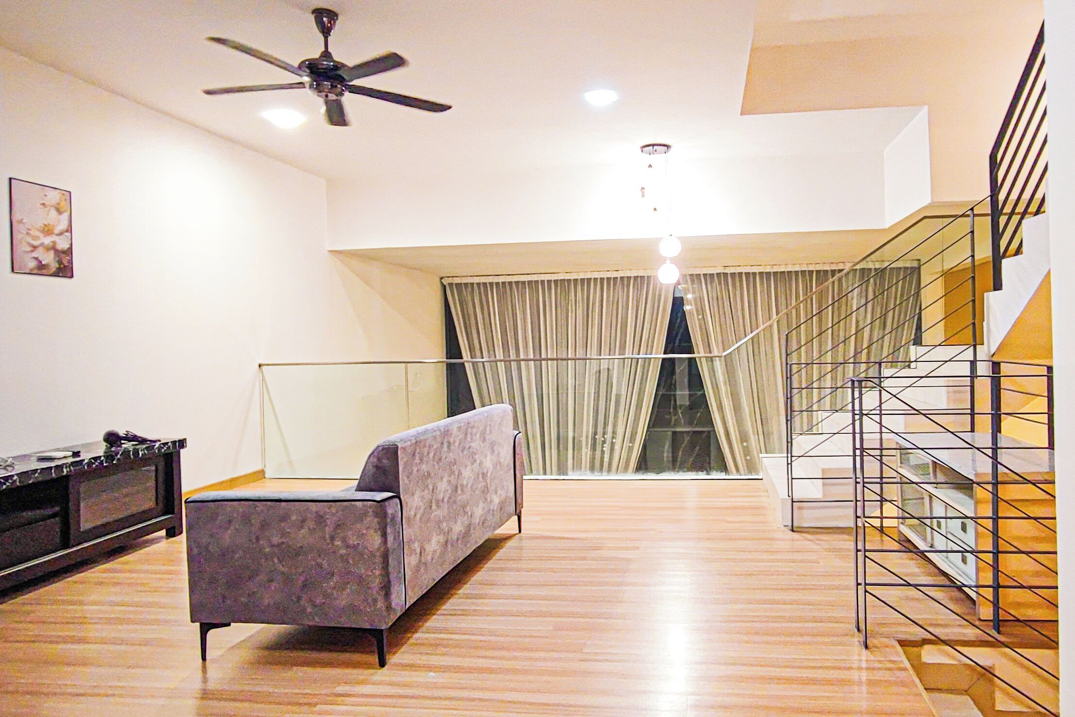 A modern living room with a sofa, a ceiling fan, a staircase, and a view through a window with curtains.