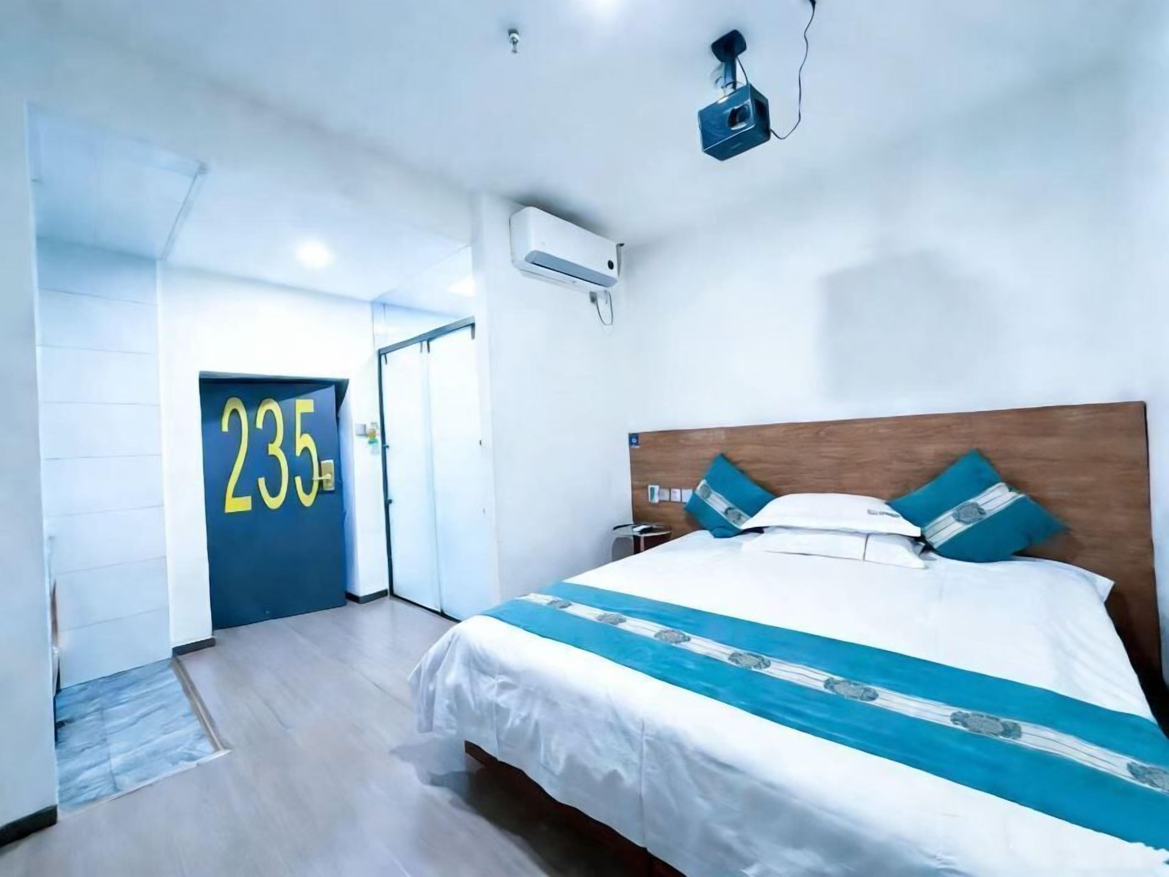 A hotel room with a bed, a door marked '235', an air conditioning unit, and a projector on the ceiling.