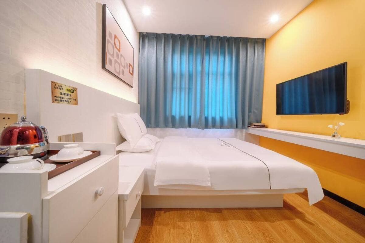 A hotel room with a bed, a desk with a kettle, a flat-screen TV, and a window with curtains.