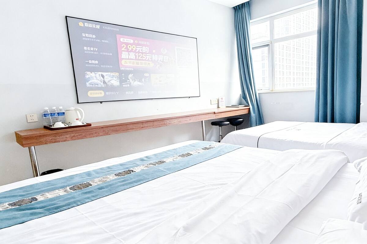 A hotel room with two beds, a desk with a computer, a window with blue curtains, and a wall-mounted TV displaying content.