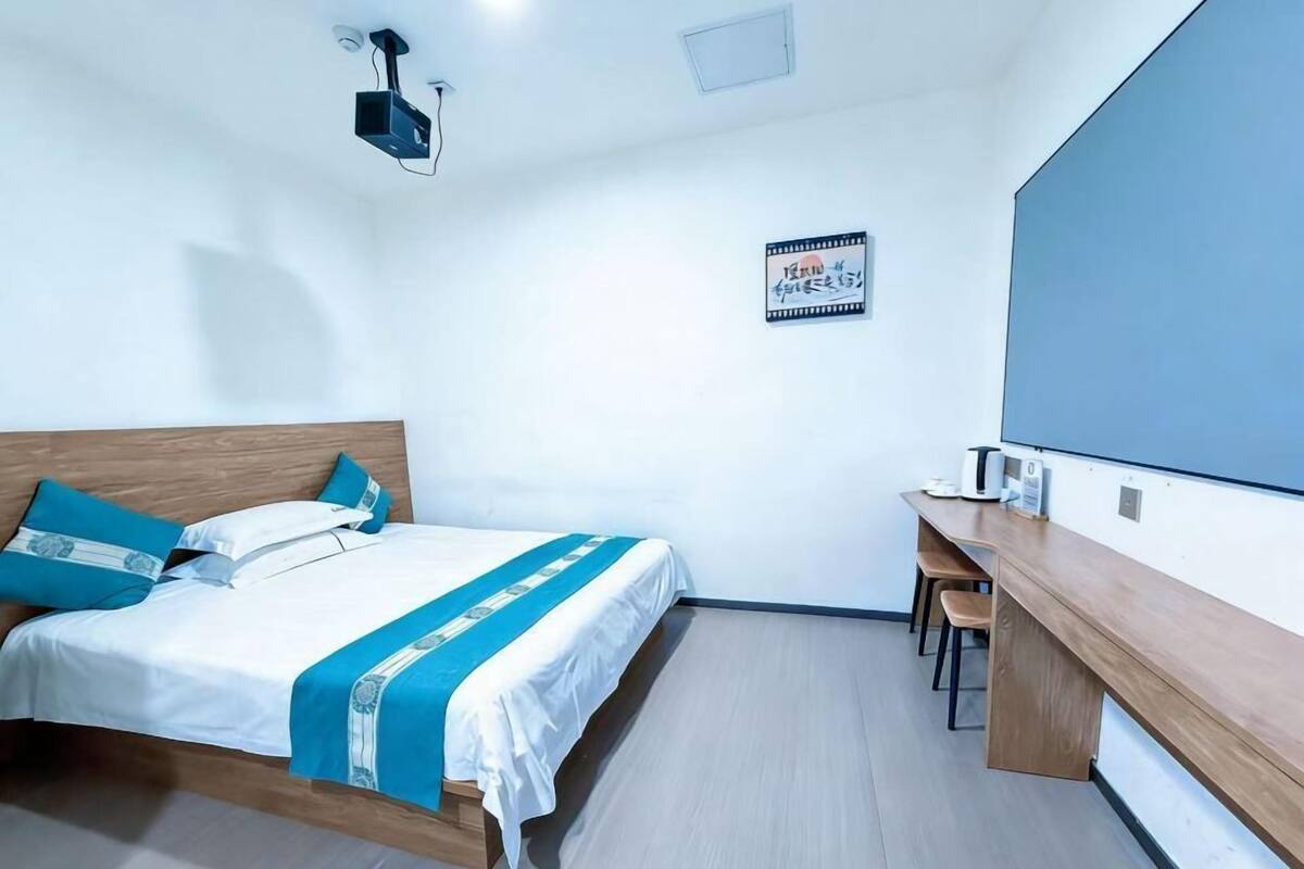 A hotel room with a bed, a desk, a chair, a projector, and a wall-mounted screen.