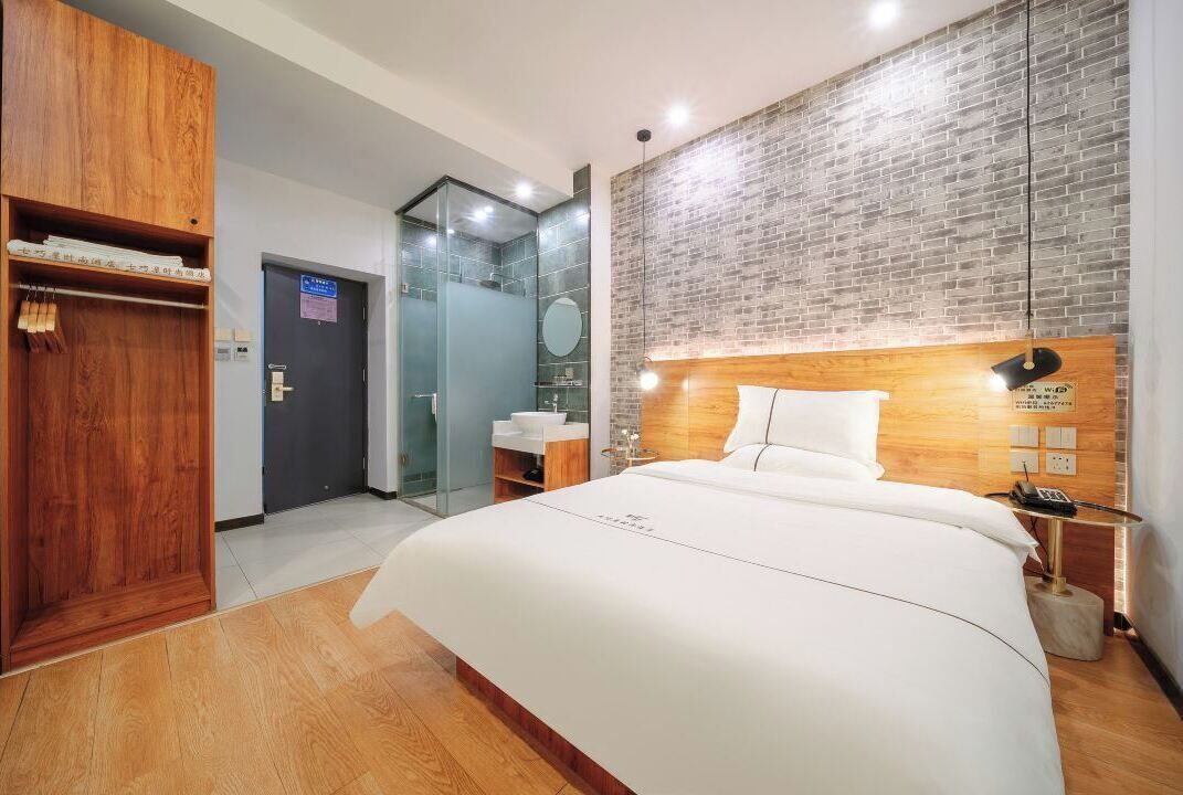 A hotel room with a large bed, wooden wardrobe, a glass shower enclosure, and a brick-patterned wall.