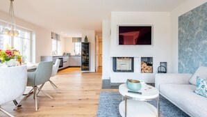 A modern living room with a sofa, a dining area with chairs, a kitchen in the background, a fireplace, and a TV mounted on the wall. - Sjipwai-Hüs 13c - Sjipwai-Hüs 13c in Westerland (Sylt)
