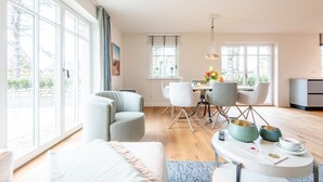 A modern living room with a sofa, a dining area with chairs, a kitchen area, and large windows providing natural light. - Sjipwai-Hüs 13c - Sjipwai-Hüs 13c in Westerland (Sylt)