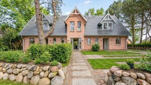 Une maison en briques avec un chemin de pierre menant à l’entrée, entourée de verdure et d’arbres.