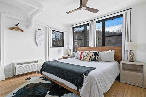Quarto com cama, duas mesas de cabeceira, ventoinha no teto, janela com cortinas e radiador.