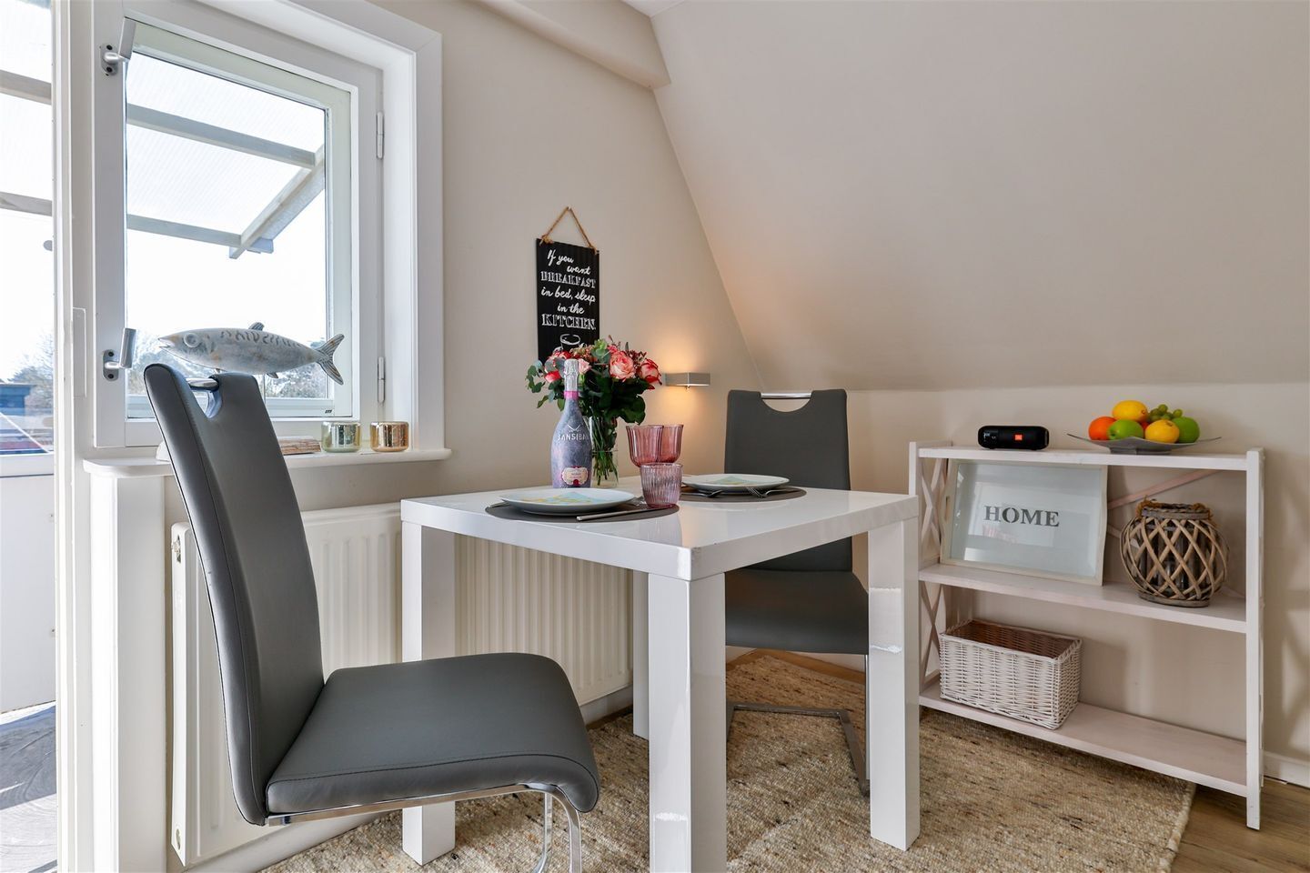 A modern kitchen with a white table, grey chairs, a hanging sign, a fruit bowl, and a 'HOME' sign on a shelf.