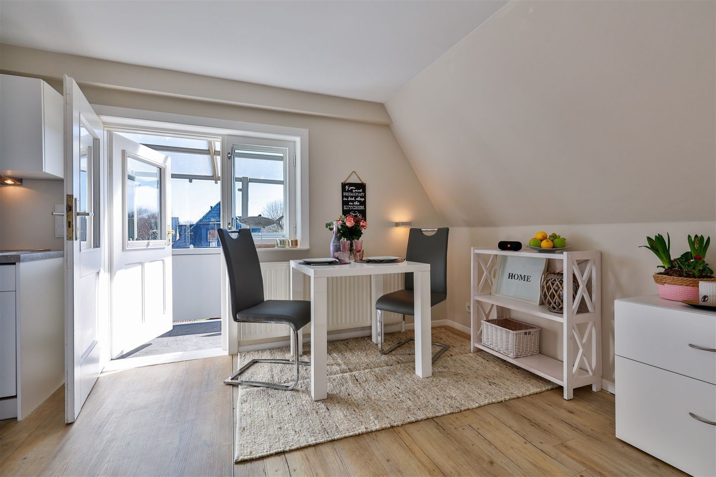 A modern kitchen with white cabinetry, a dining area with a table and chairs, a view through a large window, and a white shelf with a 'HOME' sign.