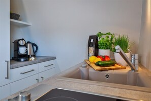 Una cocina equipada con cafetera, dispensador de pasta, verduras frescas y un lavamanos.