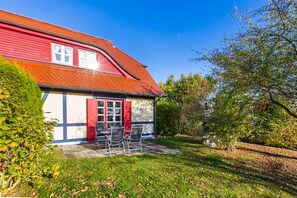 A red house with a tiled roof, a small patio with chairs, and a garden with trees and grass. - Beachfront vacation apartment with private terrace - Feriendorf Bakenberg FeWo B66 - (Dranske)