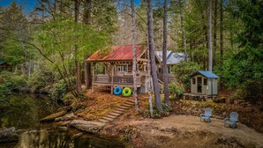 A cabin with a wooden deck, a small shed, and a dock extending into a lake. - Riverfront Cabin | Sauna • Hot Tub • Private Trail (Cashiers)