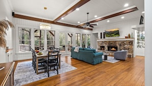 A spacious living area with a fireplace, dining table, and large windows. - Riverfront Cabin | Sauna • Hot Tub • Private Trail (Cashiers)