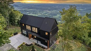 Ein zweistöckiges Haus mit dunklem Dach und zahlreichen Fenstern, umgeben von Bäumen und Blick auf ein Tal.