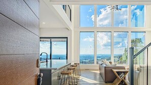 Amplia sala de estar con techos altos, ventanas grandes, una cocina con barra y vistas al exterior.