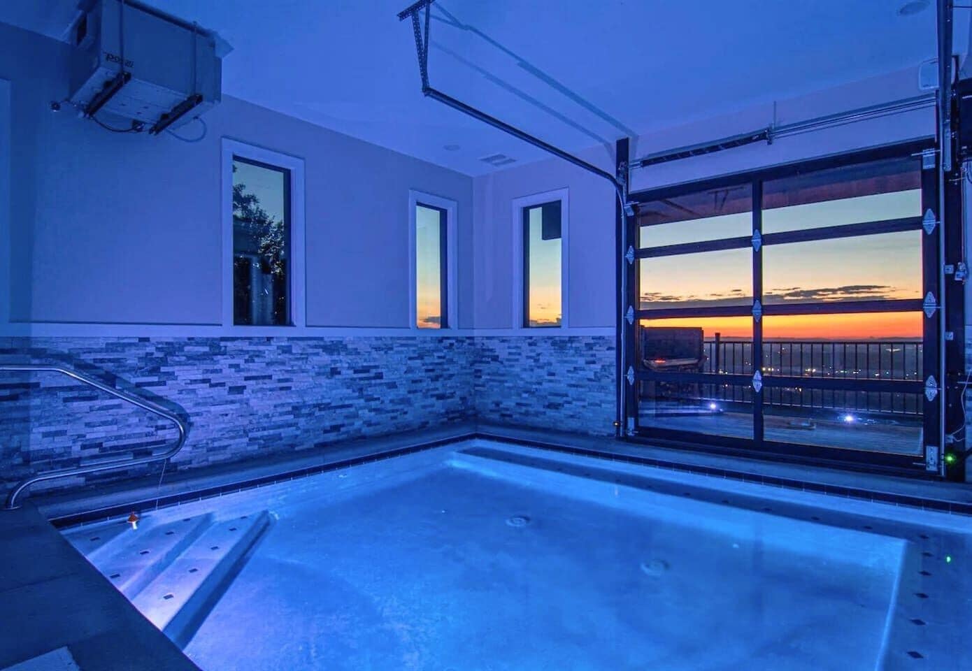 Un espace bien-être comprenant un grand jacuzzi, une porte vitrée donnant sur un balcon avec vue sur le coucher de soleil, et un mur recouvert de carreaux.