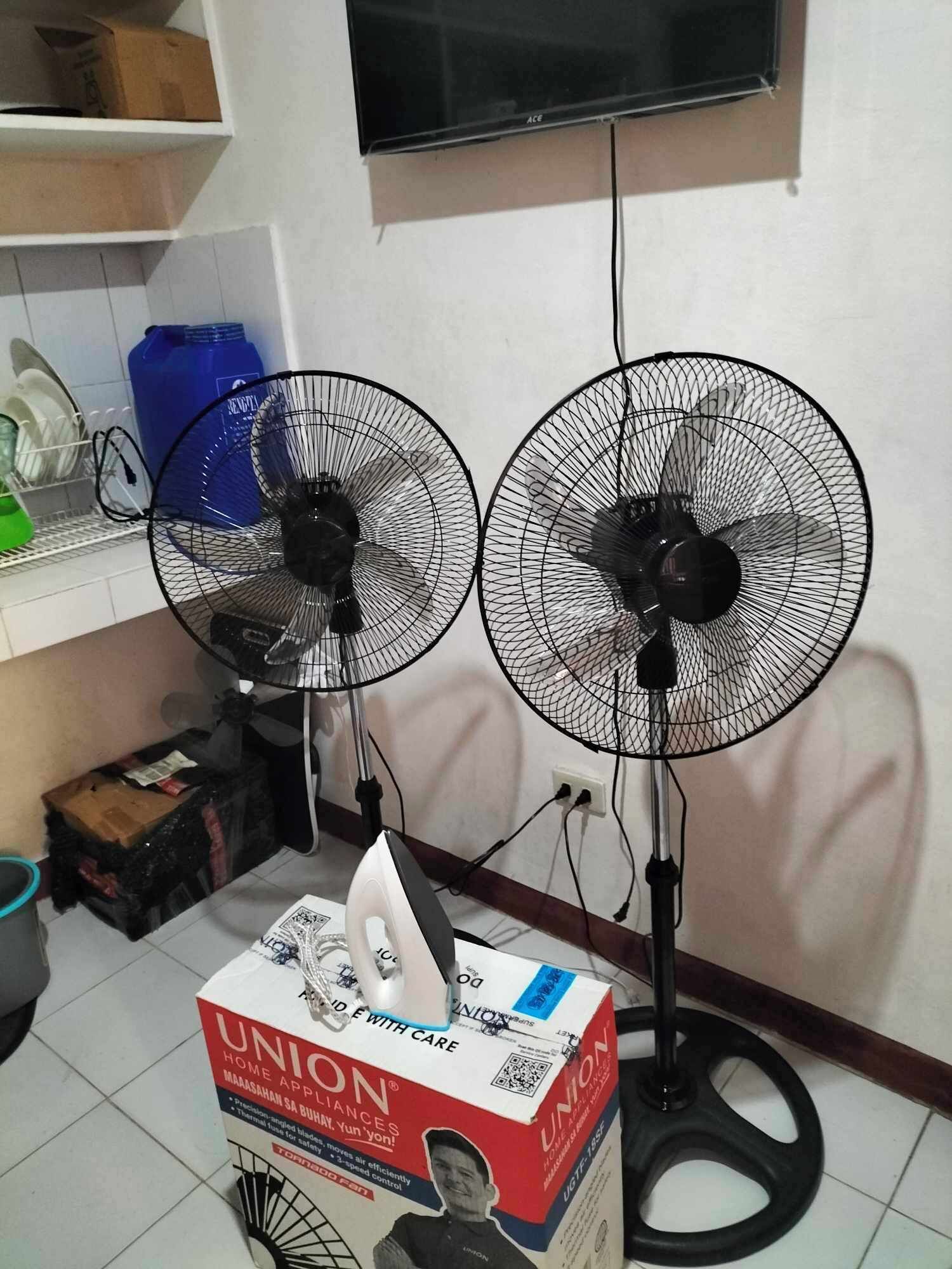 Deux ventilateurs sur pied, un téléviseur et une boîte d’appareils électroménagers de la marque Union se trouvent dans une pièce dont le sol est carrelé et qui est équipée d’une étagère où sont rangés de la vaisselle.