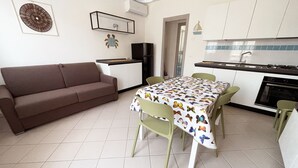 En kompakt opholdsstue med sofa, spisebord, stole, tekøkken og et lille køleskab.