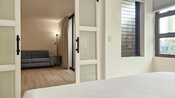 A bedroom with a bed, a grey sofa, a lamp, and a view of a building through a window. - Waikiki Banyan with Free Parking & Washer/Dryer in Unit! (Honolulu)
