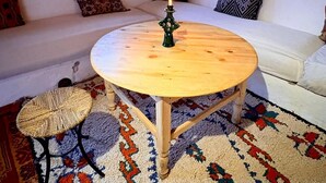 A cozy living room with a round wooden table, a lit candle, a woven stool, a patterned rug, and a white sofa. - Tigmi Hamid (Assfalou)