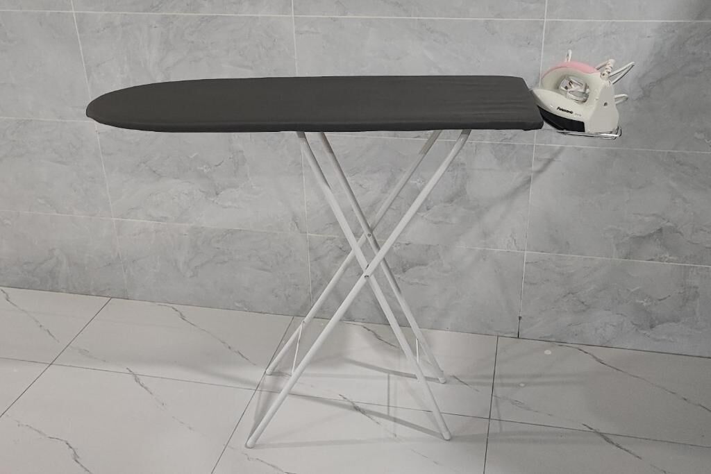 A portable ironing board with a white base and a black surface, positioned against a tiled wall.