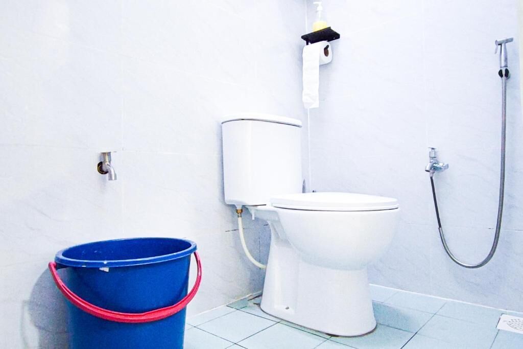 A bathroom with a white toilet, a blue bucket, a shower hose, and a toilet paper holder.