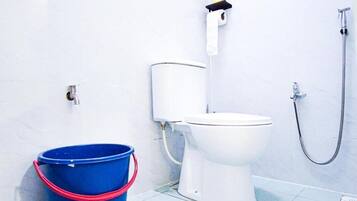 A bathroom with a white toilet, a blue bucket, a shower hose, and a toilet paper holder.