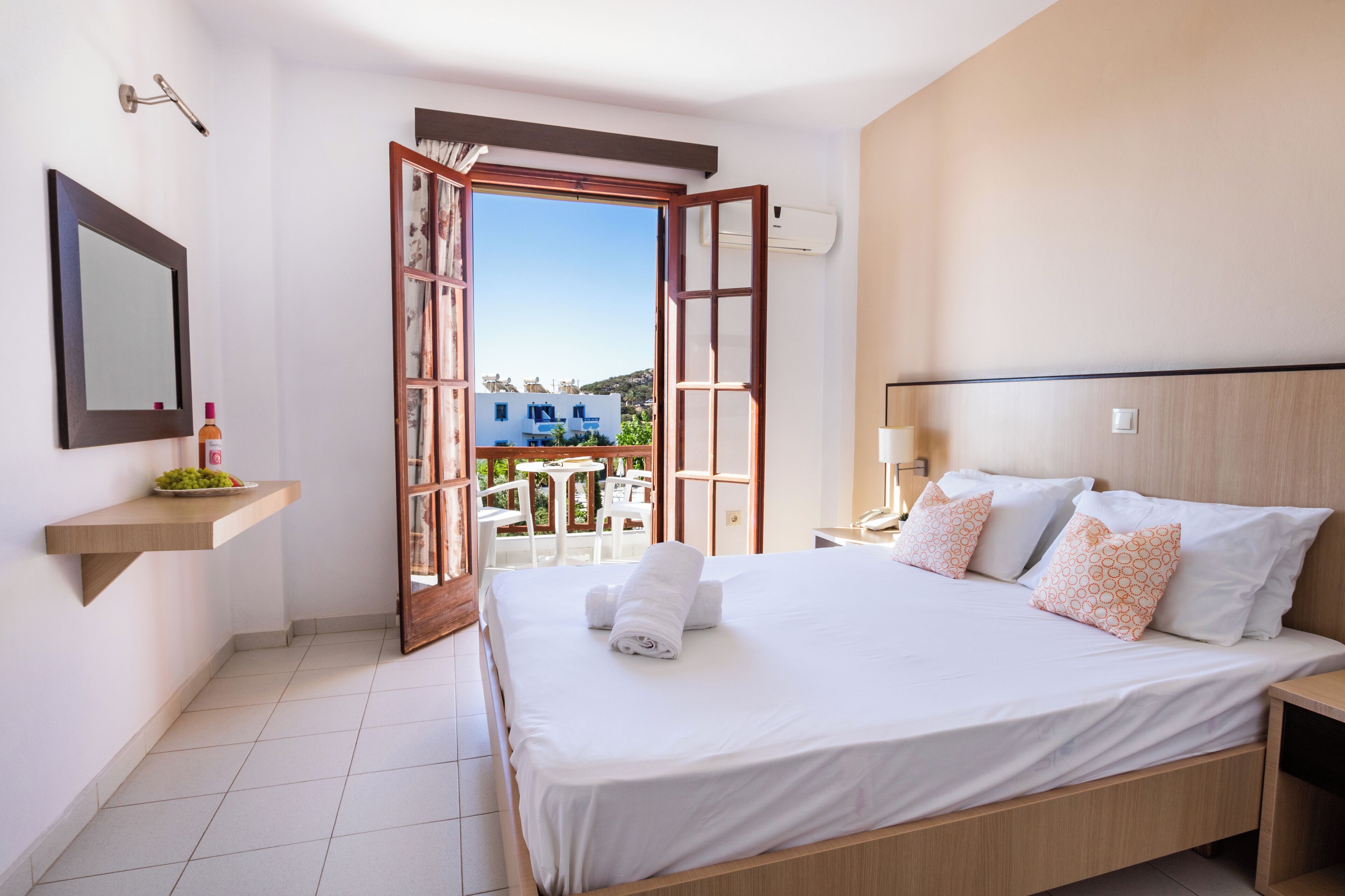 Habitación de hotel con cama, mesita de noche, espejo, estante con una botella y fruta, y puerta abierta que da a un área exterior con mesa y sillas.