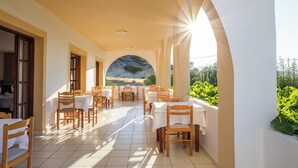A sunlit outdoor dining area with tables and chairs, a view of a rocky landscape, and a white wall. - Casa Garden (Karpathos)