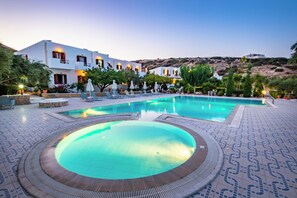 A pool area with lounge chairs, a circular hot tub, and a building with multiple balconies. - Casa Garden (Karpathos)