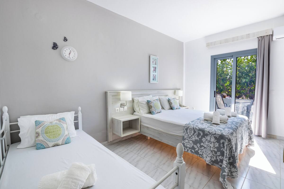 Ein Schlafzimmer mit zwei Betten, einem Nachttisch, einer Wanduhr, einem gerahmten Bild und einem Fenster mit Vorhängen.