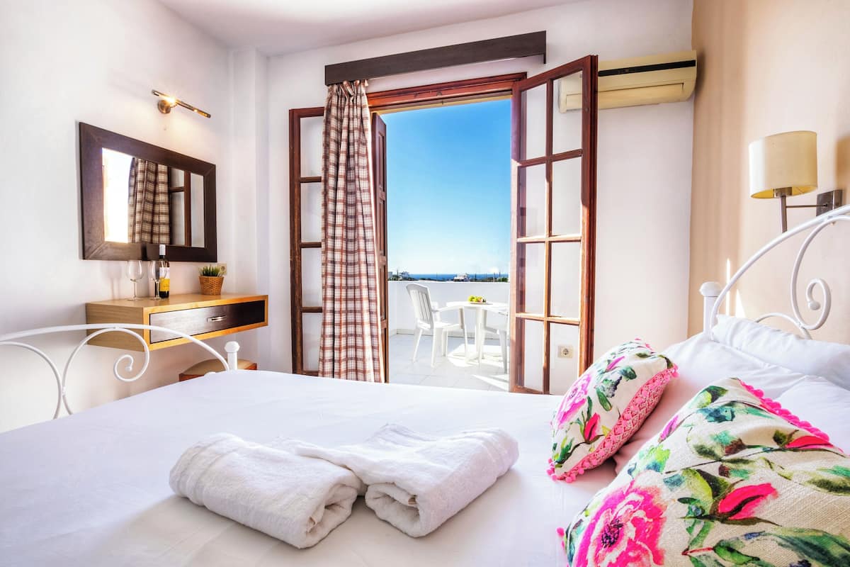 Ein Hotelzimmer mit Bett, Nachttisch mit Lampe, Spiegel und offenem Fenster mit Blick.
